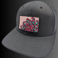 A dark gray trucker cap with a rectangular patch on the front featuring a pink and teal floral design, set against a dark gray to black gradient background.