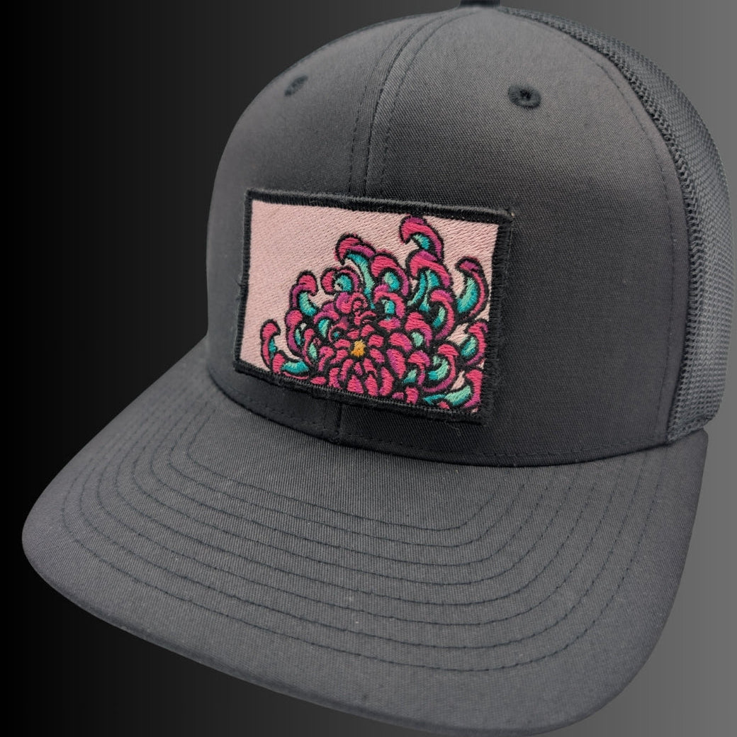 A dark gray trucker cap with a rectangular patch on the front featuring a pink and teal floral design, set against a dark gray to black gradient background.