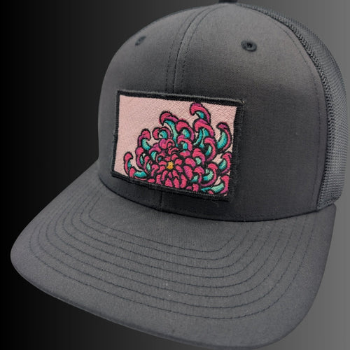 A dark gray trucker cap with a rectangular patch on the front featuring a pink and teal floral design, set against a dark gray to black gradient background.