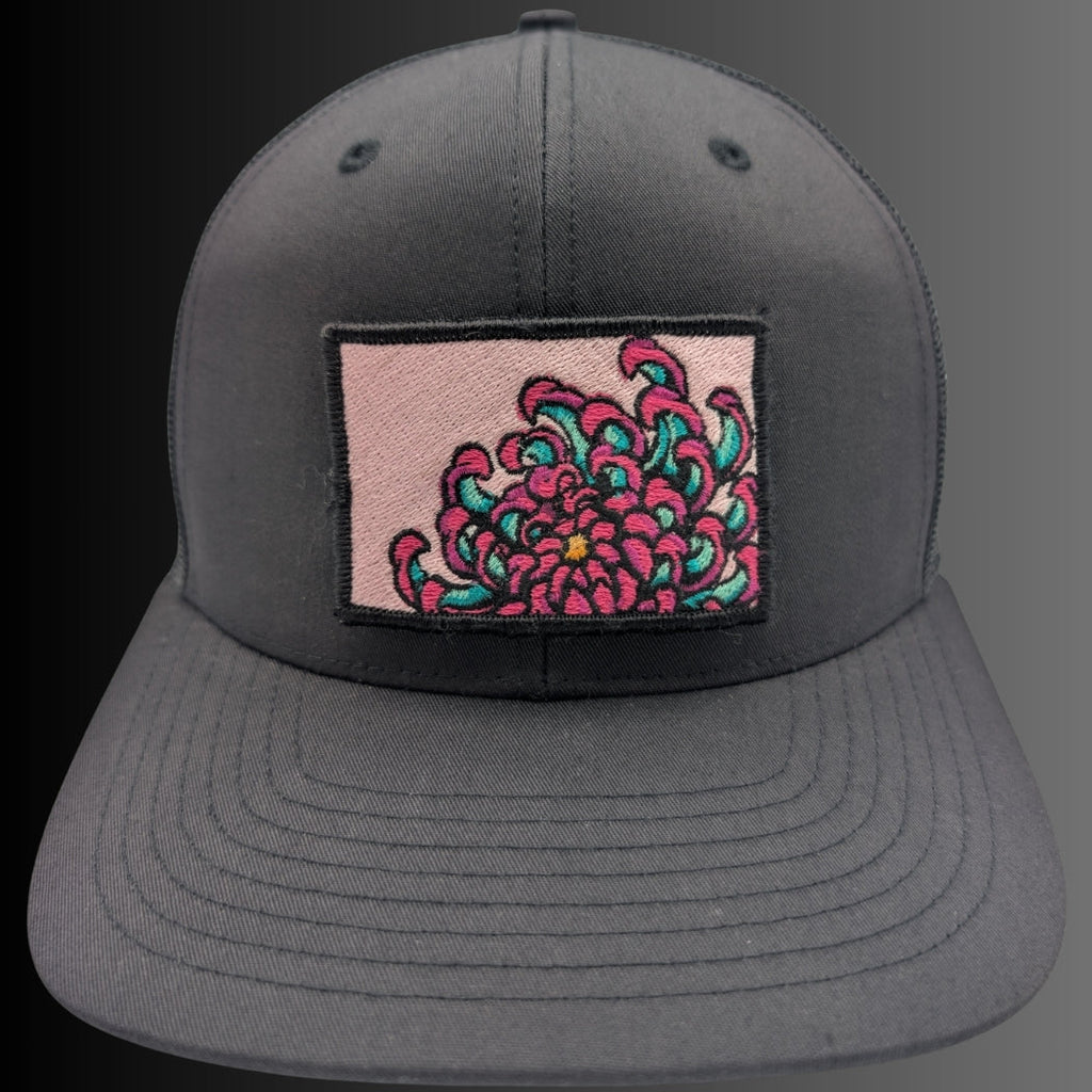 A dark gray snapback cap featuring a rectangular patch on the front with a pink background and a detailed embroidered pink and teal flower.