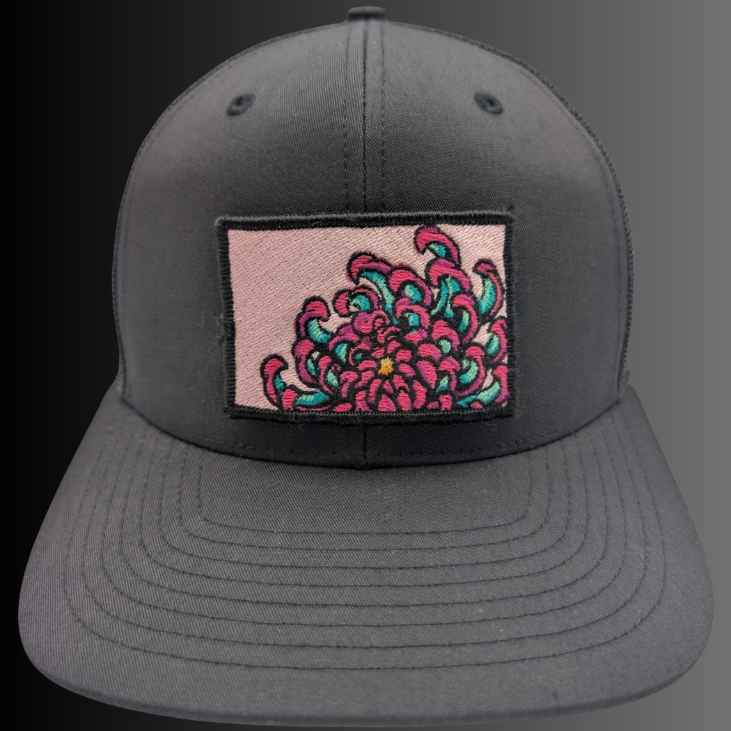 A dark gray snapback cap featuring a rectangular patch on the front with a pink background and a detailed embroidered pink and teal flower.