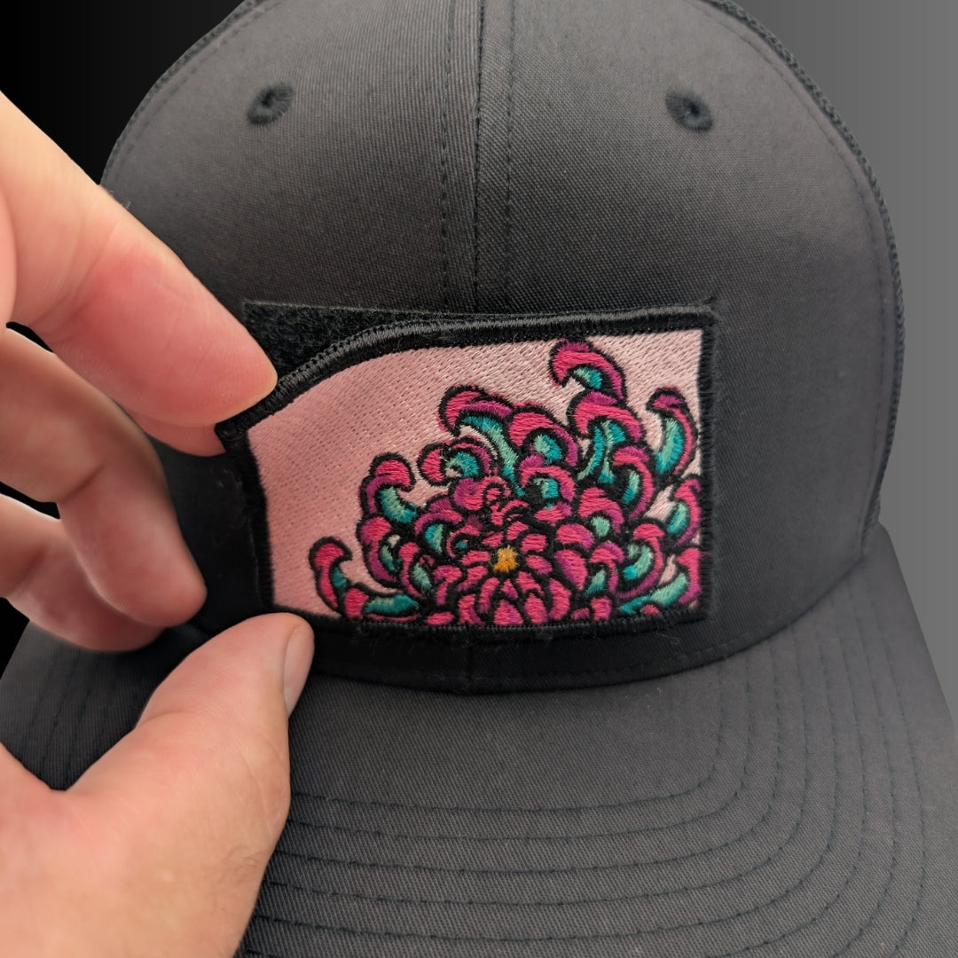 A close-up of a dark gray trucker cap with a hand holding a rectangular embroidered patch in front of the brim. The patch features a pink background with a stylized floral design in bright pink, teal, and black.