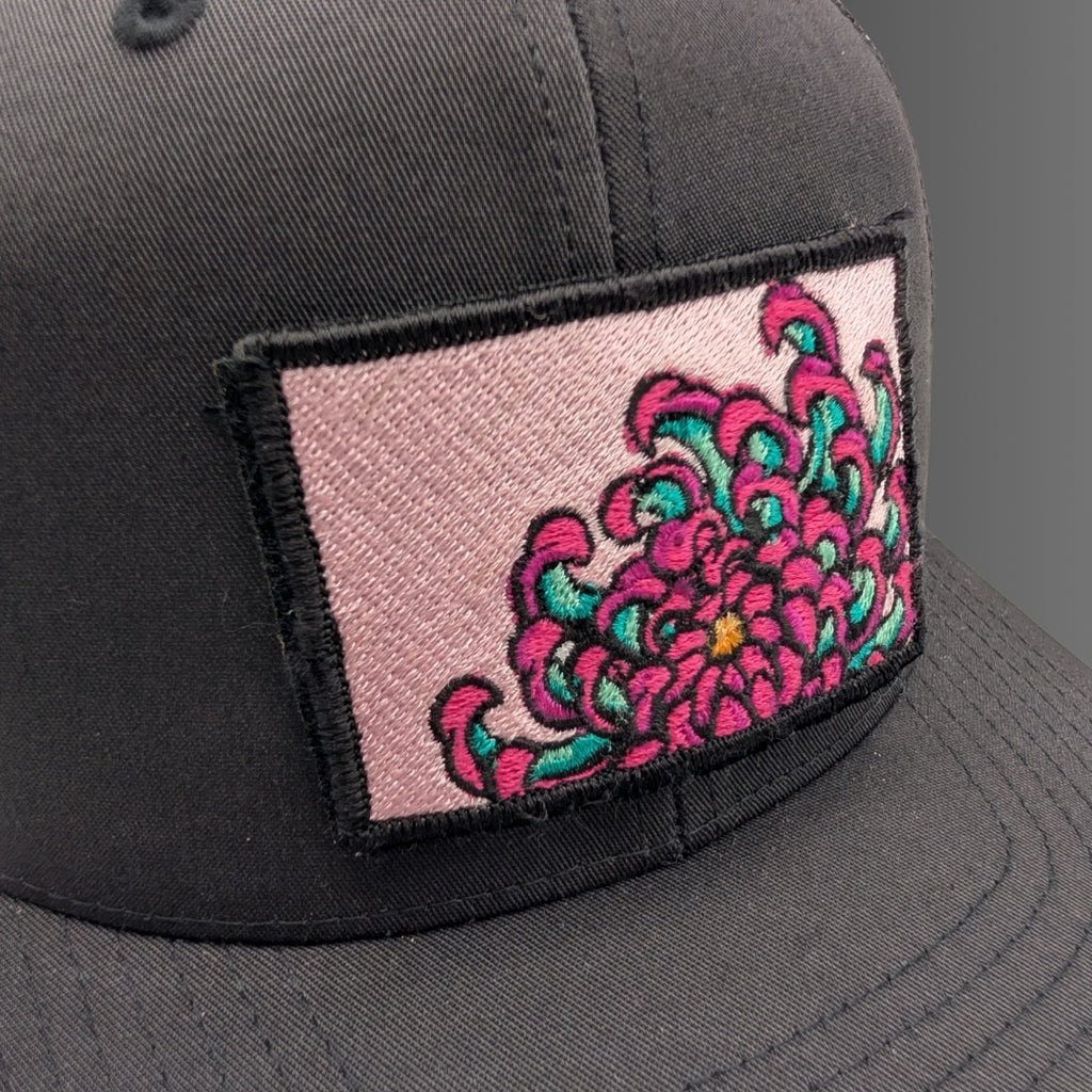 A close-up of a dark gray baseball cap featuring a rectangular embroidered patch on the front. The patch has a light pink background with a large, vibrant pink and teal floral design outlined in black.