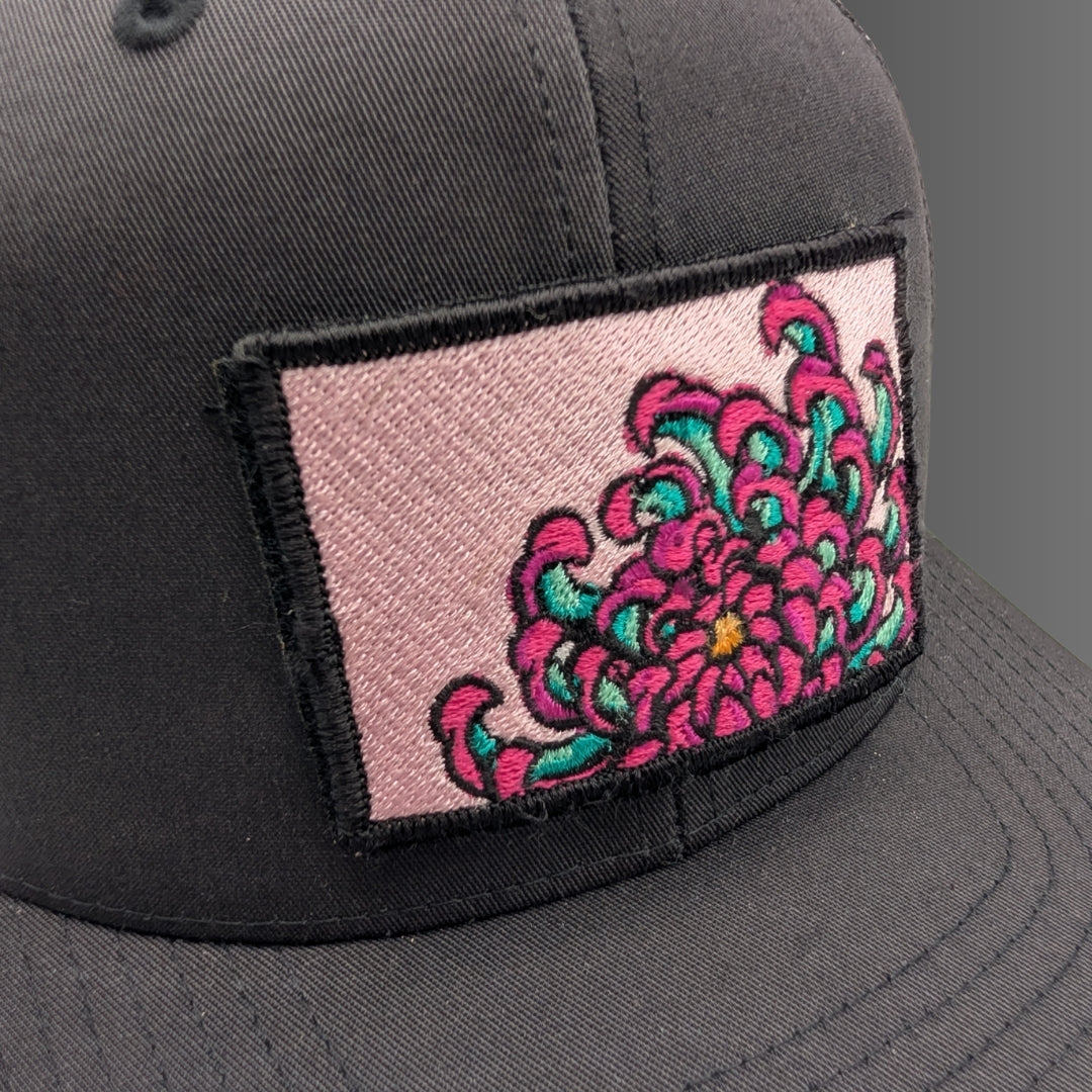 A close-up of a dark gray baseball cap featuring a rectangular embroidered patch on the front. The patch has a light pink background with a large, vibrant pink and teal floral design outlined in black.