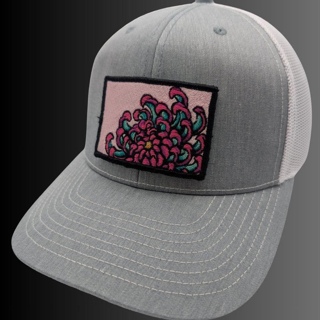 A gray trucker-style cap with white mesh back panels and a rectangular embroidered patch on the front featuring a pink and teal floral design.