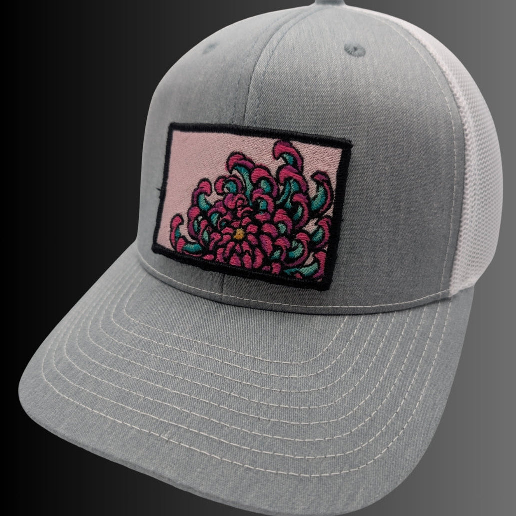 A gray trucker-style cap with white mesh back panels and a rectangular embroidered patch on the front featuring a pink and teal floral design.