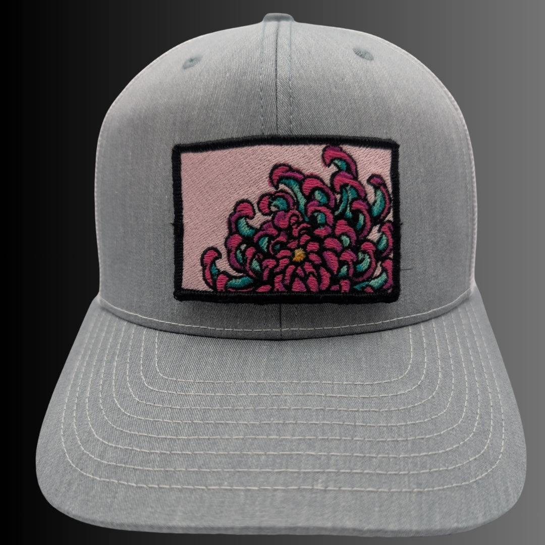 A light gray heathered baseball cap featuring a rectangular embroidered patch on the front. The patch has a light pink background with a stylized floral design in magenta and teal, outlined in black.