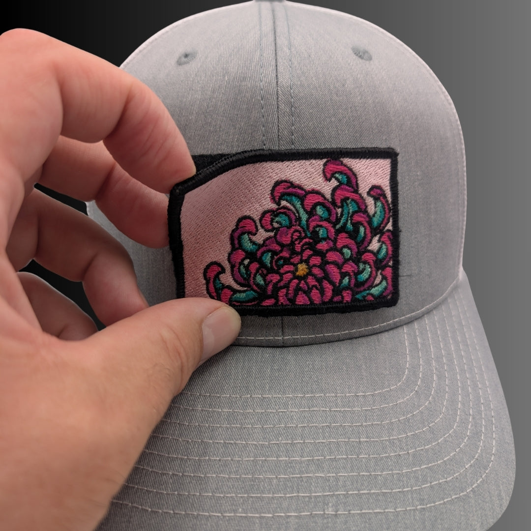 A close-up of a hand holding a gray baseball cap, showing a rectangular embroidered patch on the front with a pink background and a stylized floral design in teal and magenta with black outlines.