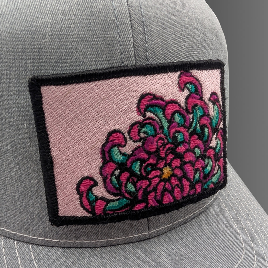 A close-up of a gray baseball cap featuring a rectangular embroidered patch on the front. The patch has a light pink background with a large stylized flower in bright pink and teal, outlined in black.