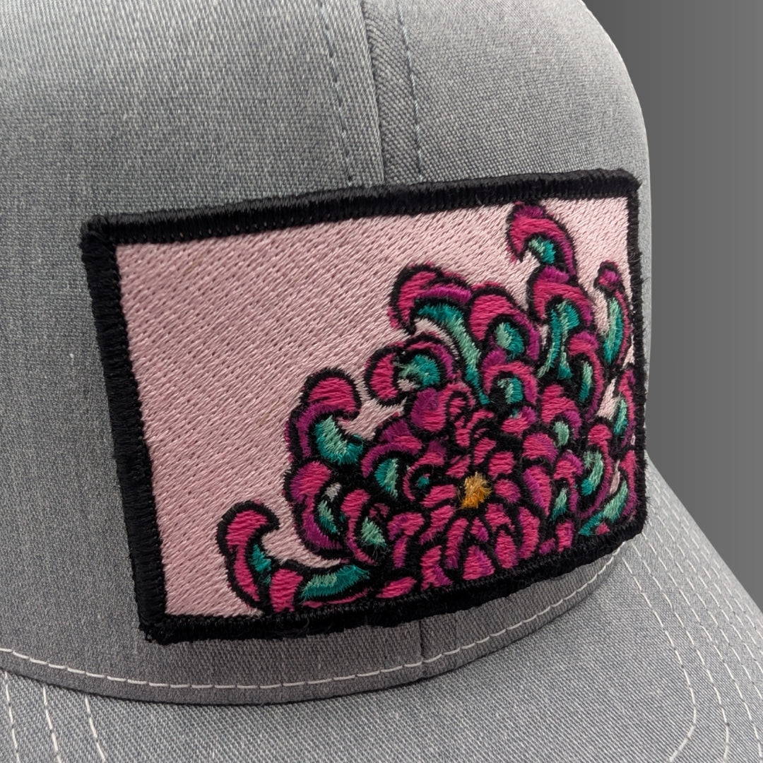 A close-up of a gray baseball cap featuring a rectangular embroidered patch on the front. The patch has a light pink background with a large stylized flower in bright pink and teal, outlined in black.