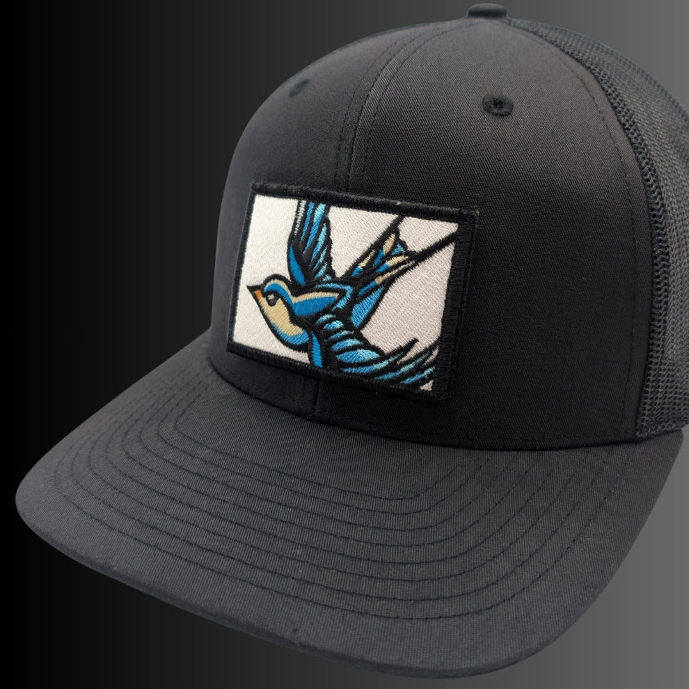 A black trucker-style baseball cap with a curved brim, black mesh back, and a rectangular embroidered patch on the front featuring a blue and white swallow in flight.