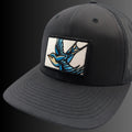 A black trucker-style baseball cap with a curved brim, black mesh back, and a rectangular embroidered patch on the front featuring a blue and white swallow in flight.