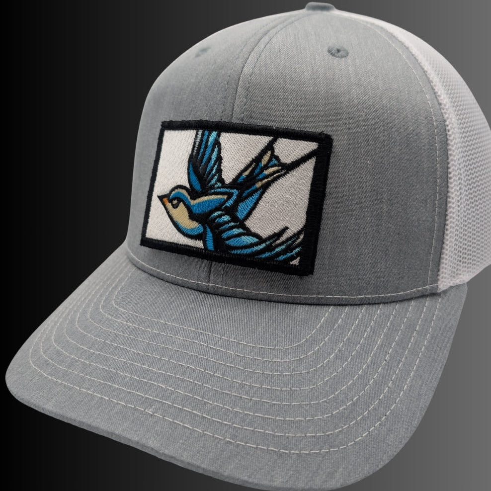 A gray trucker-style cap with a curved brim, featuring a rectangular embroidered patch on the front with a blue and tan bird design.