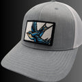 A gray trucker-style cap with a curved brim, featuring a rectangular embroidered patch on the front with a blue and tan bird design.