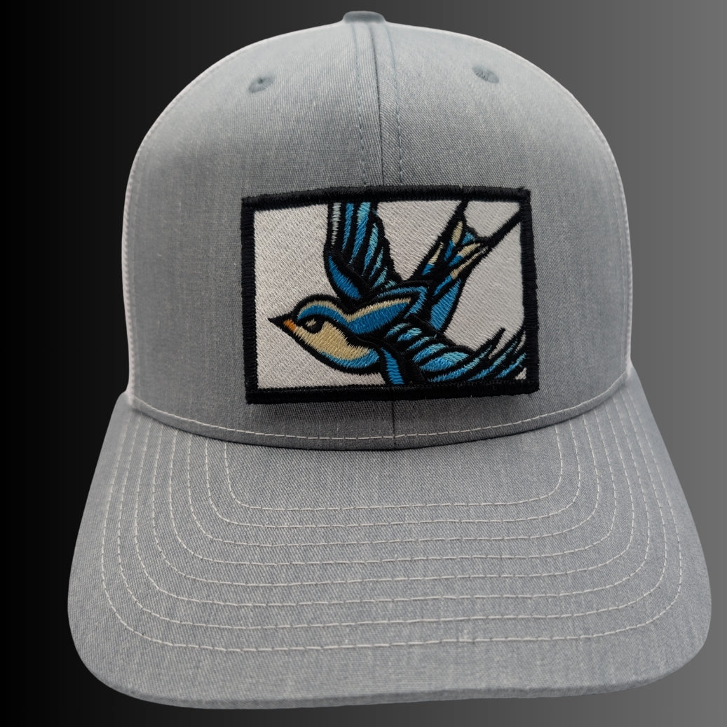 A light gray structured baseball cap with a curved brim, featuring a rectangular embroidered patch on the front with a blue and yellow bird design on a white background.