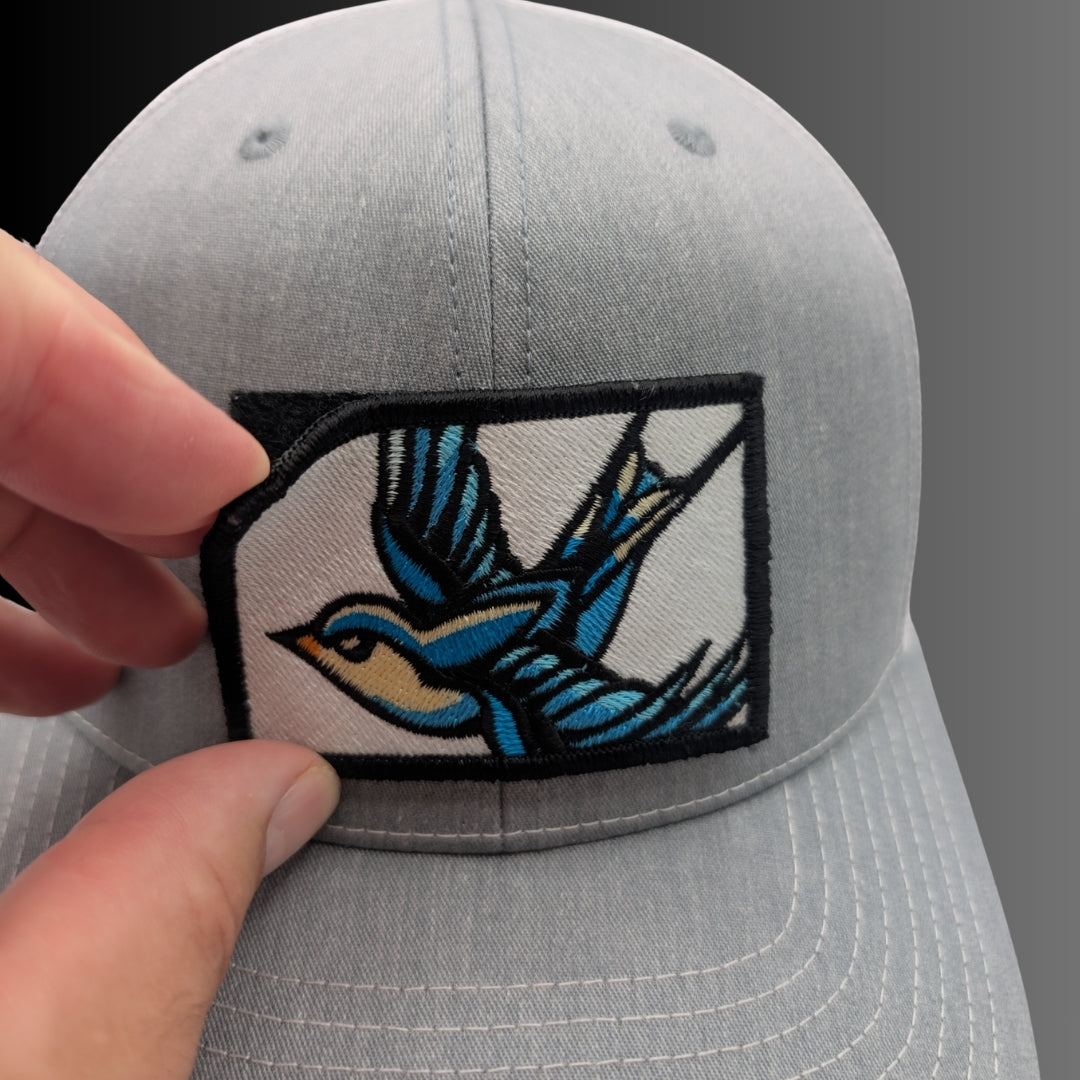 A close-up of a light gray baseball cap being held by a hand, showing a rectangular embroidered patch on the front with a stylized blue and tan bird on a white background.