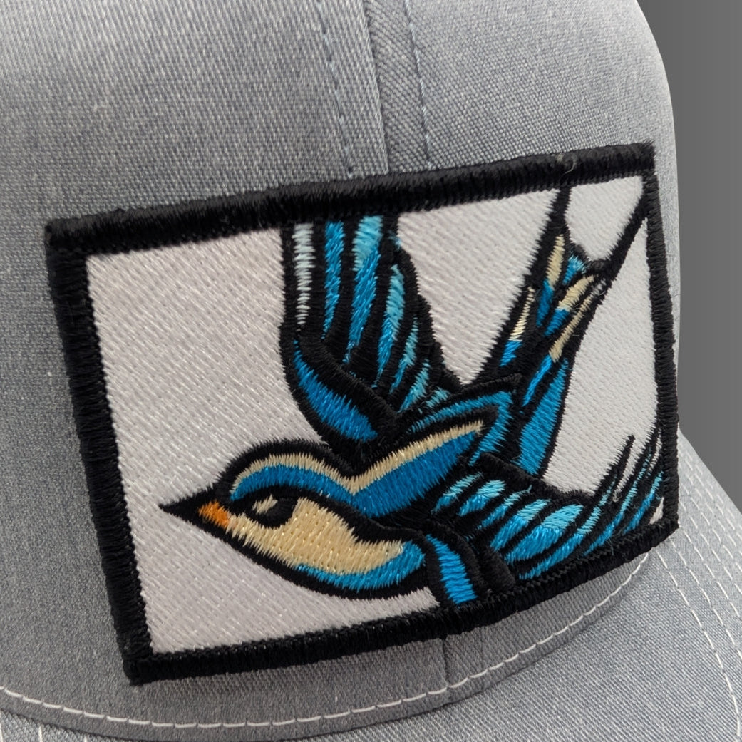 Close-up of a light gray baseball cap featuring a rectangular embroidered patch with a stylized blue and tan bird in flight outlined in black.