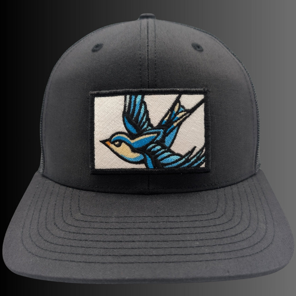 A dark gray snapback cap featuring a rectangular embroidered patch on the front with a blue and yellow swallow in flight, set against a dark gray background.